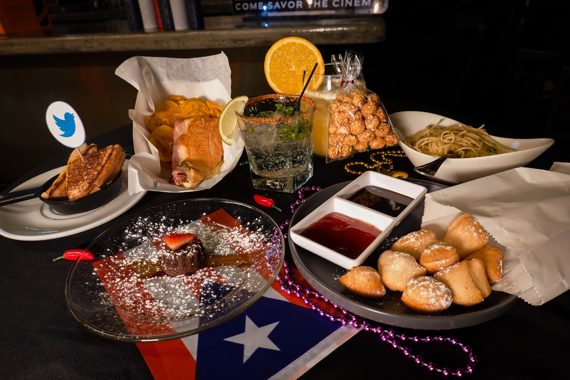 The full Chef table at Movie Thyme, shot wide: a skillet grilled cheese, a Cuban sandwich with plantain chips, a chilli-rimmed mojito, caramel popcorn, aglio e olio pasta, sugared beignets with jam and chocolate dips, and a molten cake plated over a Puerto Rican flag
