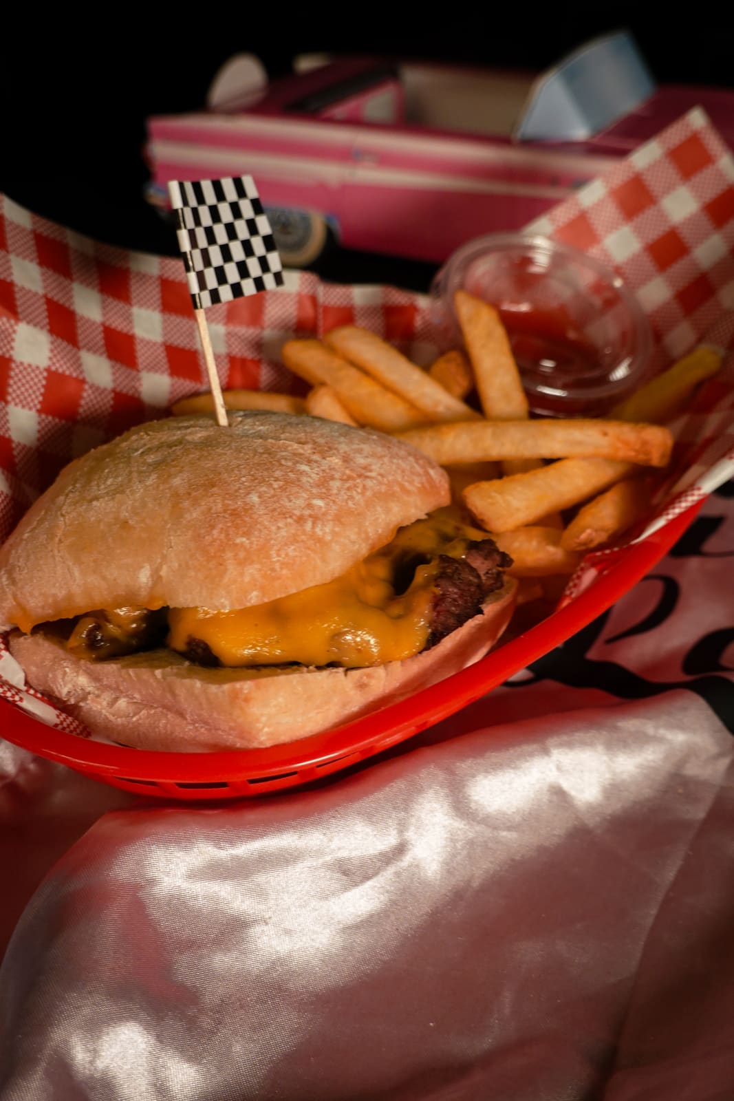 A cheeseburger and fries in a red checkered diner basket with a checkered-flag pick on pink satin