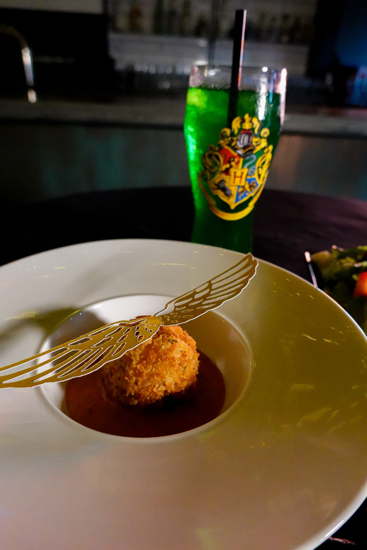 A Harry Potter course at Movie Thyme: a crisp golden risotto ball dressed with edible golden Snitch wings in a shallow bowl of tomato sauce, with a glowing green Hogwarts-crest drink behind it