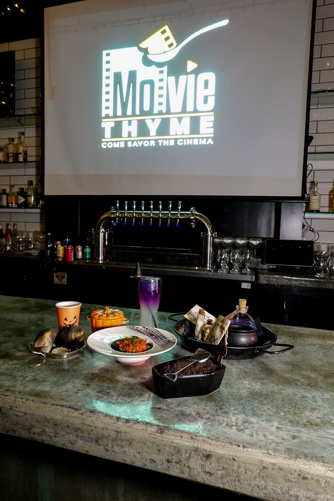 The Movie Thyme logo on a large screen above the bar and a spread of plated courses