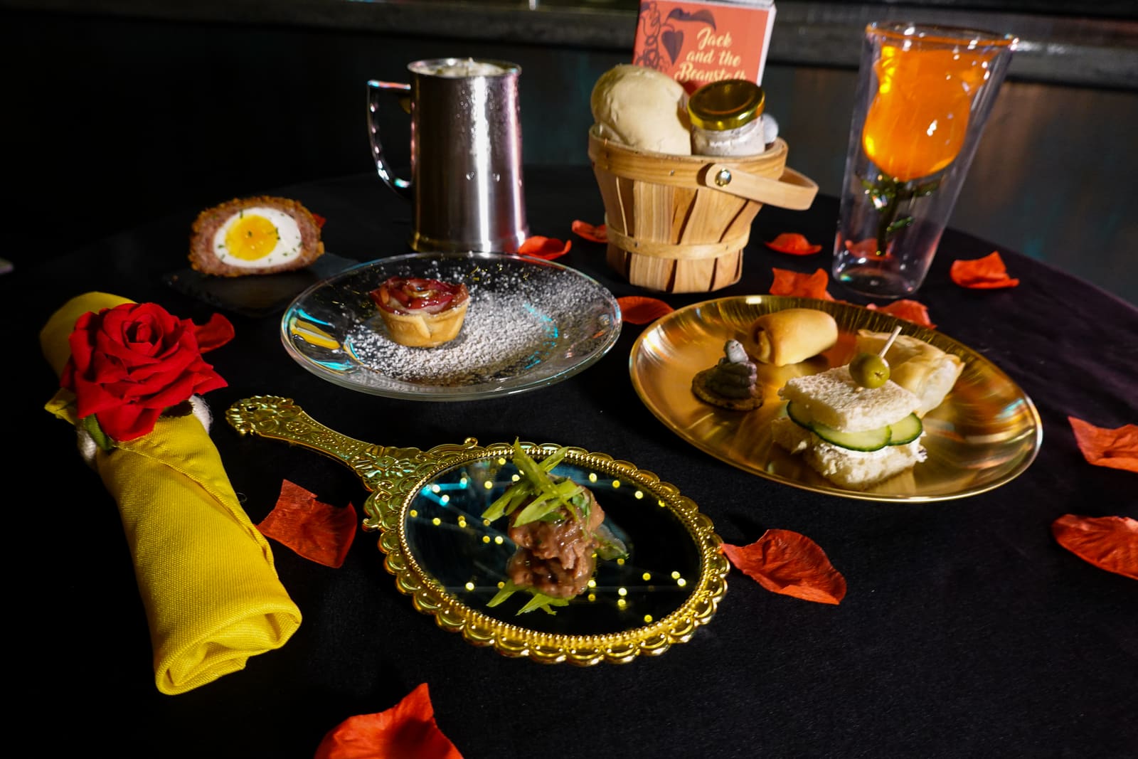 A dressed table with gold platters, red rose petals and an orange cocktail