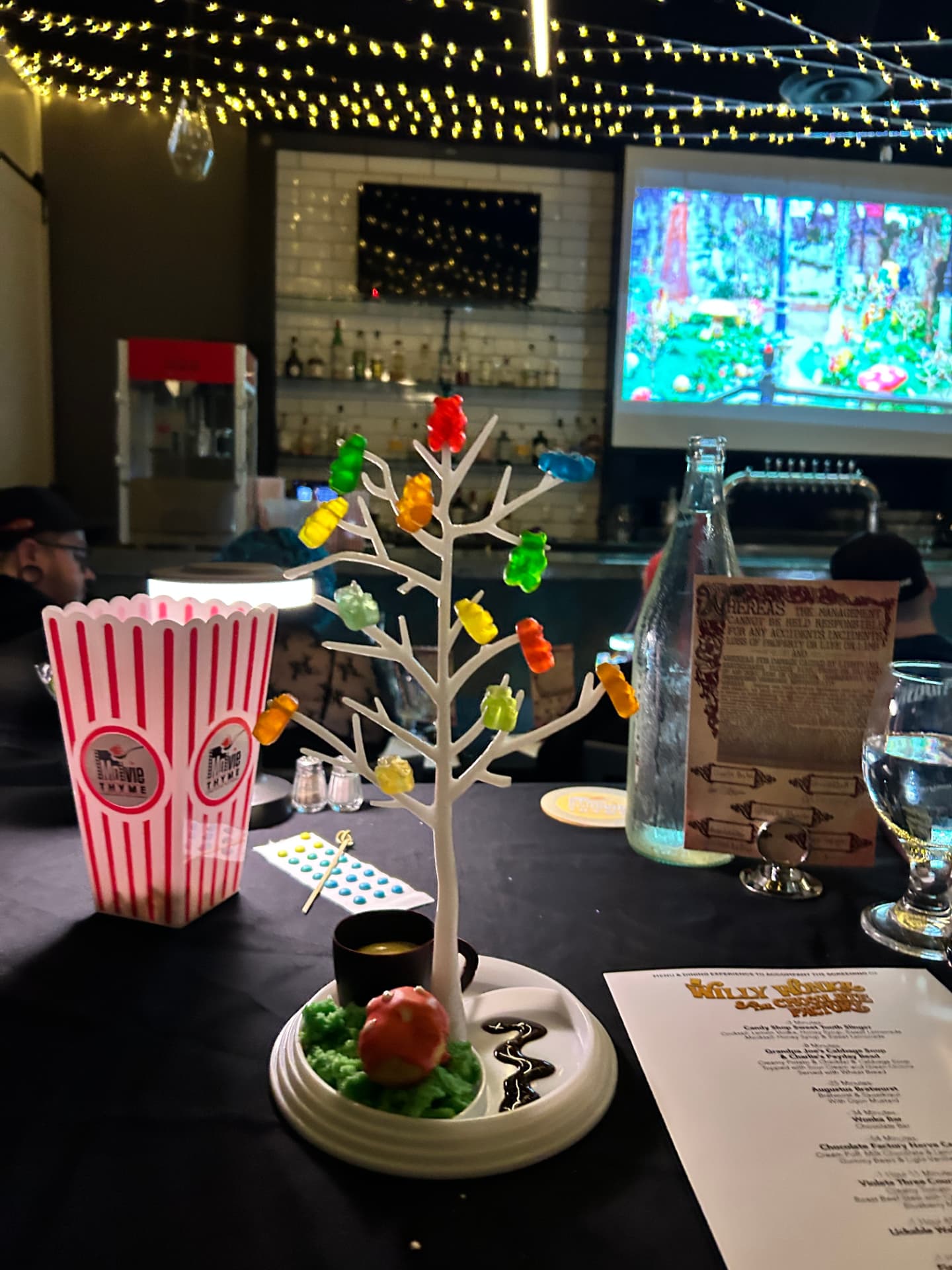 A Willy Wonka course at Movie Thyme: a white candy tree hung with gummy bears over a green 'grass' plate with a red toadstool cake, a pot of dipping chocolate and a candy button strip, beside a branded popcorn box and the printed Wonka menu, with the film playing on the screen behind under fairy lights